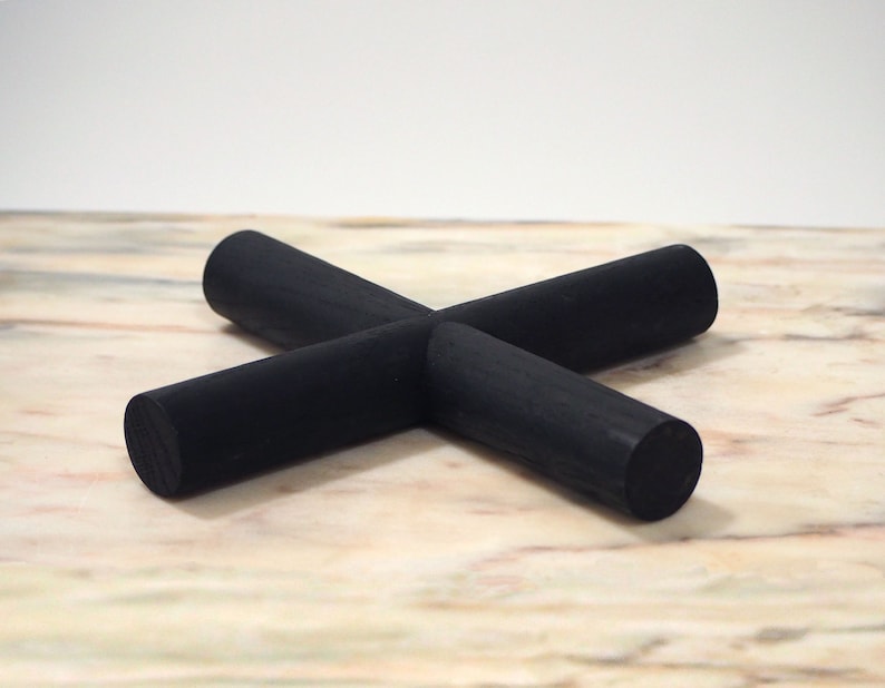May include: A black wooden coaster in a cross shape. The coaster is made of four cylindrical pieces of wood that intersect in the centre. The coaster is on a light-coloured surface.