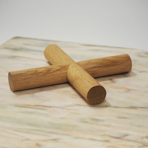 May include: A wooden trivet made from four intersecting cylindrical pieces. The trivet is constructed from light-coloured wood and arranged in a cross shape. It rests on a marble surface with beige, white, and pink tones.