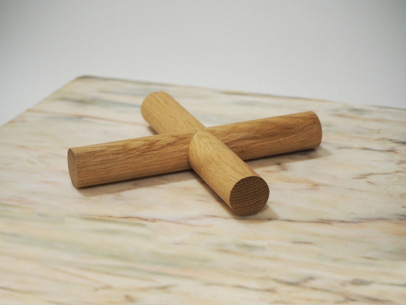 May include: A wooden trivet made from four intersecting cylindrical pieces. The trivet is constructed from light-coloured wood and arranged in a cross shape. It rests on a marble surface with beige, white, and pink tones.