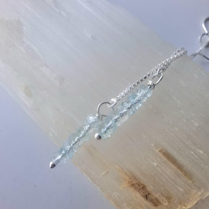 May include: A delicate necklace featuring a strand of light blue aquamarine beads. The beads are strung on a silver chain, with a small silver bead accent. The necklace is displayed on a light-coloured, textured surface.