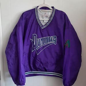 May include: A purple Diamondbacks pullover jacket with a V-neck collar. The jacket features the team's logo on the front and a patch on the sleeve. The collar and cuffs have green and white stripes. The jacket is hanging on a white hanger.