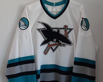 May include: White hockey jersey with teal and black stripes. The front of the jersey features a teal, black, and orange San Jose Sharks logo with a shark and two hockey sticks. The sleeves have a teal and white wave logo.