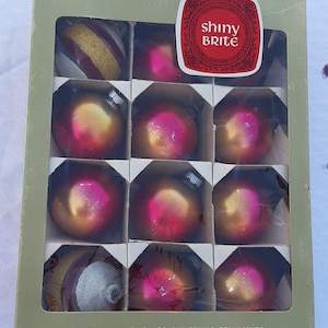 May include: A box of Shiny Brite Christmas ornaments. The box contains twelve glass ornaments in a variety of colors, including pink, gold, and red. The box is green and has the words "Finest Quality Glass Christmas Ornaments" printed on it.