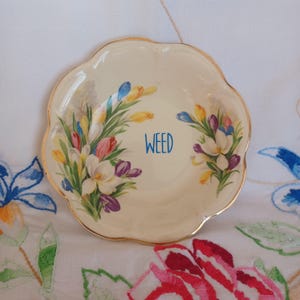 May include: A cream-coloured, scalloped-edge dish with a gold rim. The dish is decorated with colourful floral bouquets and the word "WEED" in blue script. The flowers include white, purple, yellow, and blue blooms.