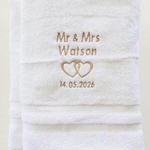 Pu&ograve; includere: Asciugamani da bagno bianchi ricamati con "Mr & Mrs Watson", due cuori intrecciati e la data 14.05.2026 in oro. Gli asciugamani sono piegati e presentano una superficie strutturata con fasce decorative.