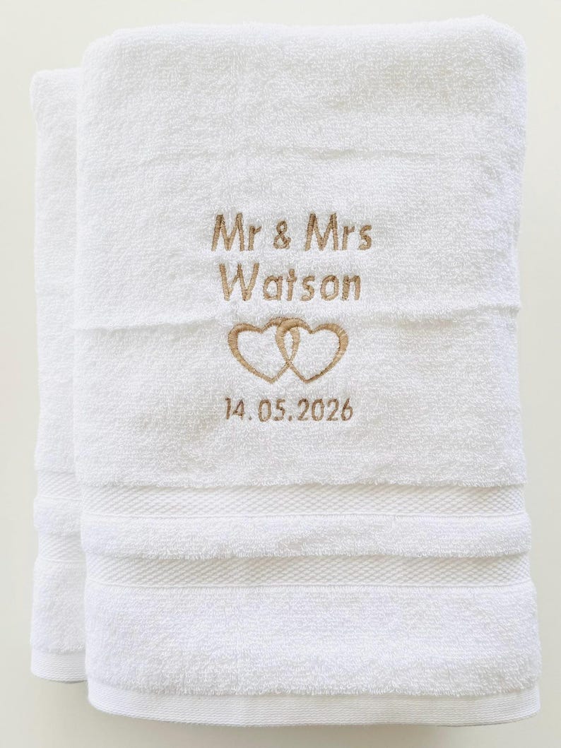 Pu&ograve; includere: Asciugamani da bagno bianchi ricamati con "Mr & Mrs Watson", due cuori intrecciati e la data 14.05.2026 in oro. Gli asciugamani sono piegati e presentano una superficie strutturata con fasce decorative.