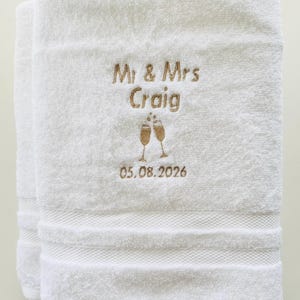 Pu&ograve; includere: Asciugamani bianchi ricamati con "Mr & Mrs Craig" in filo dorato, due calici di champagne e la data 05.08.2026. Gli asciugamani hanno un bordo strutturato.