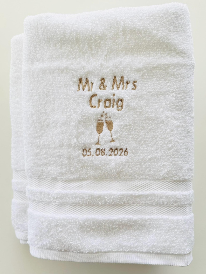 Pu&ograve; includere: Asciugamani bianchi ricamati con "Mr & Mrs Craig" in filo dorato, due calici di champagne e la data 05.08.2026. Gli asciugamani hanno un bordo strutturato.