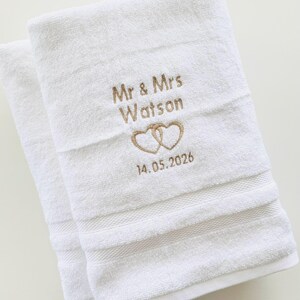 Pu&ograve; includere: Due asciugamani bianchi con ricamo "Mr & Mrs Watson" sopra due cuori intrecciati e la data "14.05.2026". Gli asciugamani hanno una superficie strutturata e strisce orizzontali decorative.