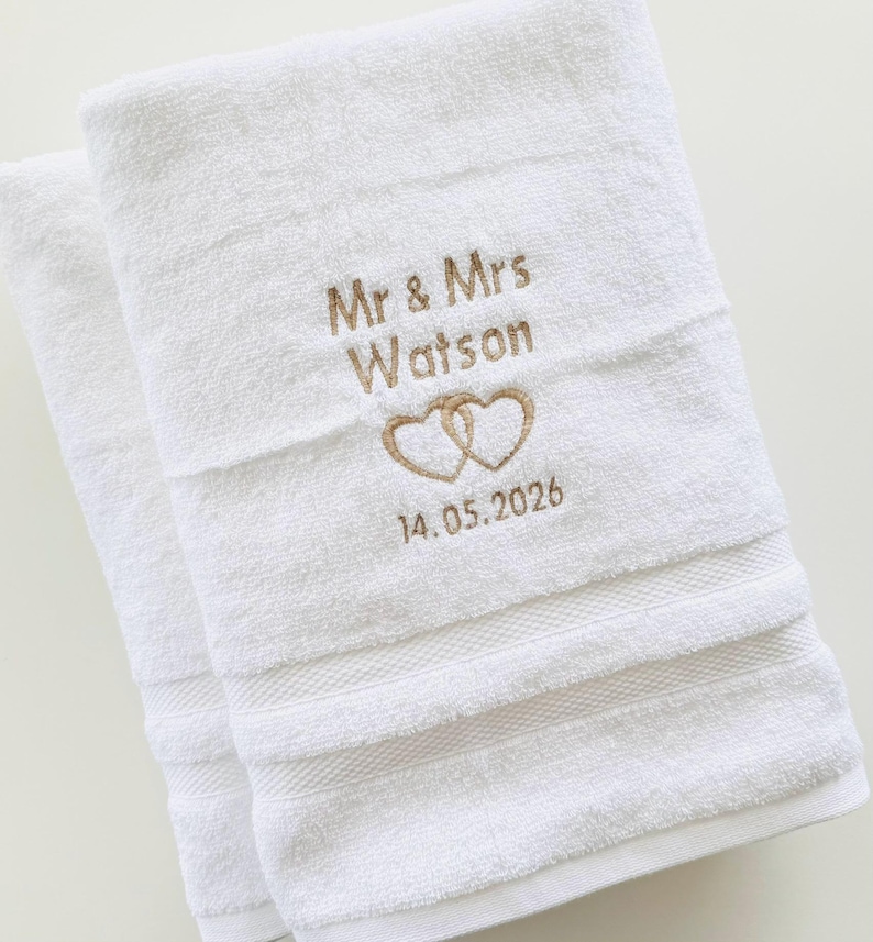 Pu&ograve; includere: Due asciugamani bianchi con ricamo "Mr & Mrs Watson" sopra due cuori intrecciati e la data "14.05.2026". Gli asciugamani hanno una superficie strutturata e strisce orizzontali decorative.