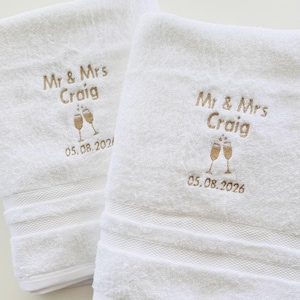 Pu&ograve; includere: Due asciugamani bianchi ricamati con "Mr & Mrs Craig", due calici di champagne e la data "05.08.2026" in filo dorato. Gli asciugamani hanno un bordo strutturato.