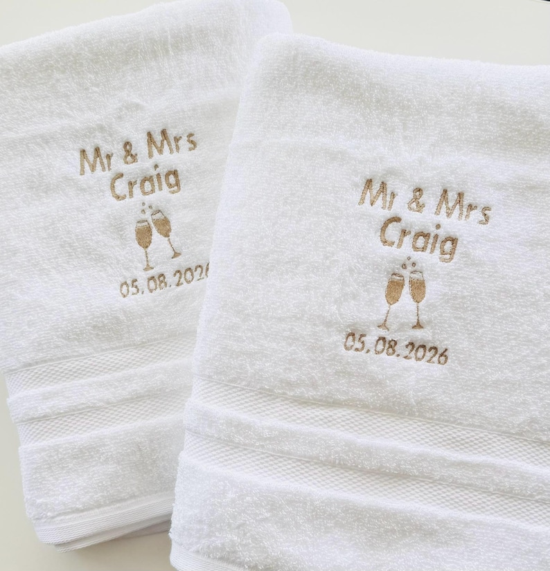 Pu&ograve; includere: Due asciugamani bianchi ricamati con "Mr & Mrs Craig", due calici di champagne e la data "05.08.2026" in filo dorato. Gli asciugamani hanno un bordo strutturato.