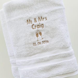 Pu&ograve; includere: Due asciugamani bianchi piegati con ricamo dorato. Il testo recita "Mr & Mrs Craig" sopra la data "05.08.2026", con calici di champagne. Gli asciugamani hanno un bordo strutturato.