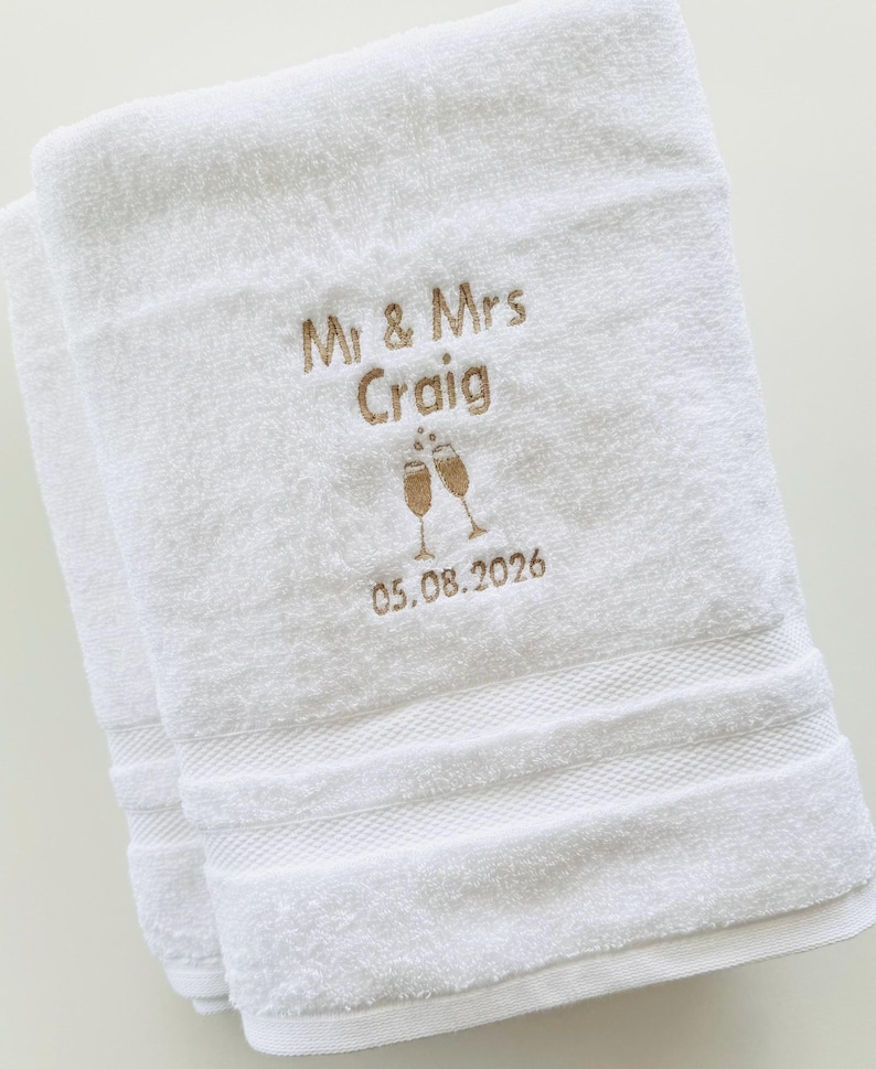 Pu&ograve; includere: Due asciugamani bianchi piegati con ricamo dorato. Il testo recita "Mr & Mrs Craig" sopra la data "05.08.2026", con calici di champagne. Gli asciugamani hanno un bordo strutturato.