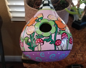 May include: A hand-painted gourd birdhouse with a pink background, green leaves, orange and yellow flowers, and red and white mushrooms. The birdhouse has a checkered black and white roof and a purple base with green and white polka dots.