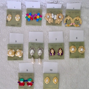 Puede incluir: Una colección de pendientes vintage, cada uno exhibido en una tarjeta verde. Los estilos incluyen pendientes de clip dorados, con perlas y adornos de joyas. Las tarjetas dicen "TWO SISTERS" e incluyen la palabra "WHIMSES".