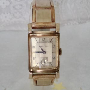 May include: A vintage gold-coloured Waltham wristwatch with a rectangular face. The watch has a cream-coloured dial with black numerals and hands. The watch strap is also gold-coloured, with a classic link design. A classic timepiece.