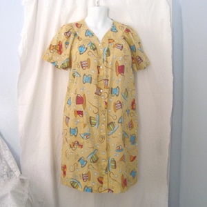 Può includere: Un accappatoio giallo con bottoni e maniche corte, decorato con un motivo colorato di rocchetti di filo, tazze e altre immagini legate al cucito. L'accappatoio è esposto su un manichino su sfondo bianco.