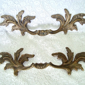 May include: Two ornate, antique brass drawer pulls with a floral leaf design. The handles are curved with detailed, symmetrical leaf patterns. The pulls are set against a white lace background.