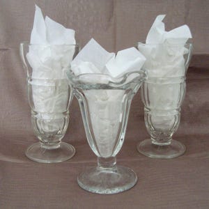Peut inclure: Trois coupes à glace en verre transparent de conception classique. Chaque verre est rempli de papier de soie blanc. Les verres ont une base ronde, un pied et un bord évasé. L'arrière-plan est de couleur unie et neutre.