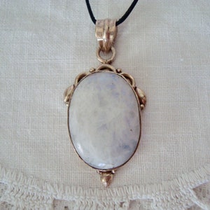 May include: A silver-toned pendant with an oval, iridescent moonstone. The stone is set in a decorative frame and hangs from a black cord. The pendant has a vintage or antique aesthetic.