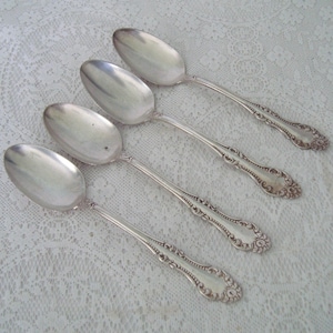 May include: Four silver spoons with ornate handles are arranged diagonally on a white lace tablecloth. The spoons have a reflective surface and a detailed floral pattern on the handles. The spoons are likely for serving.