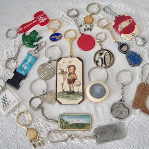 May include: A collection of vintage keychains in various shapes and designs. Includes a red frog, a blue and white ribbon, a Hard Rock Hollywood keychain, and a keychain with a picture of a boy with a birdcage. The keychains are on a white lace surface.