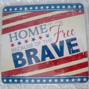 May include: Square coaster with a patriotic design. The coaster features red and white stripes, blue stars, and the text "HOME OF THE FREE BECAUSE OF THE BRAVE" in blue and red lettering. The coaster has a distressed, vintage look.