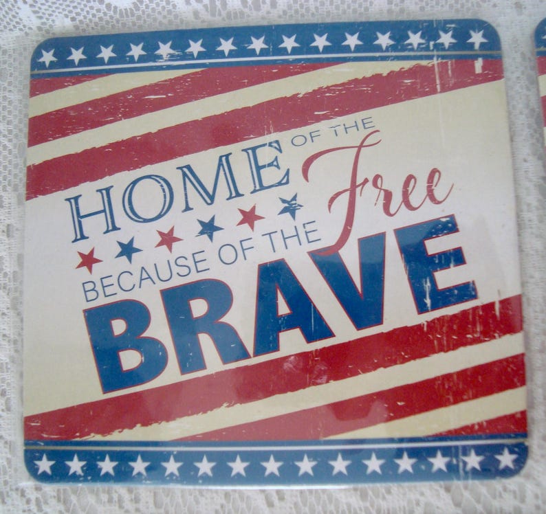 May include: Square coaster with a patriotic design. The coaster features red and white stripes, blue stars, and the text "HOME OF THE FREE BECAUSE OF THE BRAVE" in blue and red lettering. The coaster has a distressed, vintage look.