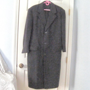 May include: A long, dark charcoal gray overcoat featuring a notched lapel collar, long sleeves, and a three-button front closure. The coat has a straight, structured silhouette with a subtle, repeating tonal pattern throughout the fabric.