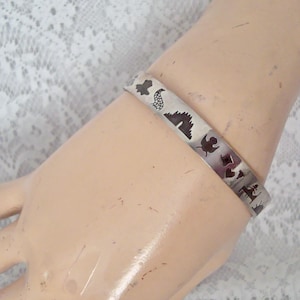 Può includere: Un bracciale rigido in metallo color argento con una serie di sagome scure tagliate al laser. I disegni includono un castello, un uccello, un cuore, una forma a diamante e un sole, disposti in un motivo ripetuto sulla superficie del bracciale.