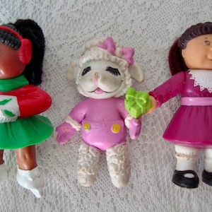 May include: Three vintage plastic dolls. One doll is wearing a red and green outfit with white gloves and shoes. The second doll is a lamb in a pink outfit. The third doll is wearing a pink dress and black shoes.