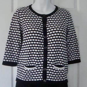 Könnte beinhalten: Eine schwarz-weiße Strickjacke mit einem sich wiederholenden ziegelartigen Muster, einem Rundhalsausschnitt, Dreiviertelärmeln, einer Knopfleiste auf der Vorderseite und zwei kleinen Taschen an der Taille. Das Kleidungsstück hat einfarbige schwarze Ränder.