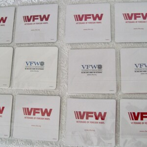 May include: A collection of white square coasters featuring the VFW logo in red or blue. The text "VETERANS OF FOREIGN WARS" is printed below the logo. Some coasters also include the text "NO ONE DOES MORE FOR VETERANS."