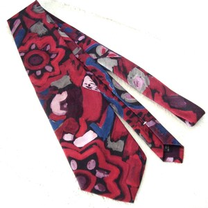 May include: A red necktie with an abstract pattern in shades of red, blue, gray, and purple. The tie features a repeating design of organic shapes and lines, creating a bold and artistic look.