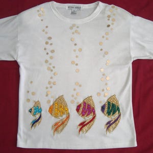 Op de afbeelding: Wit T-shirt met ronde hals met vier kleurrijke visapplicaties. Elke vis heeft een met kralen bezette kop en staart, met gouden paillettenbellen die opstijgen. Het shirt is van het merk Victoria Harbour.
