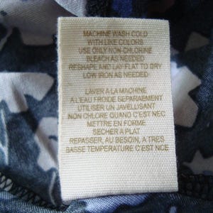 May include: Close-up of a fabric label with care instructions. The tag is beige with black text, including washing instructions such as "Machine wash cold with like colours" and "Low iron as needed."