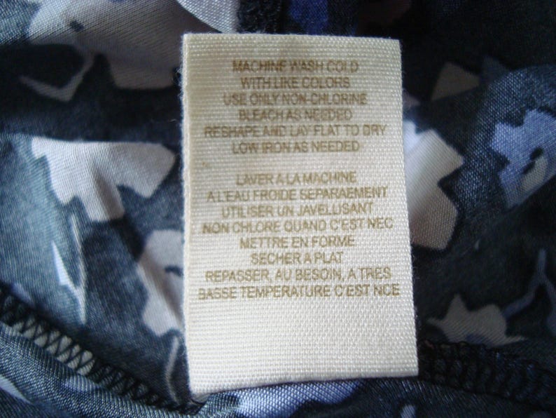 May include: Close-up of a fabric label with care instructions. The tag is beige with black text, including washing instructions such as "Machine wash cold with like colours" and "Low iron as needed."