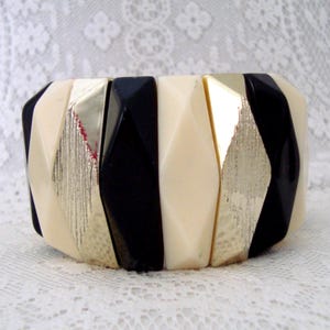 May include: A chunky bracelet with a geometric design. It features alternating segments of black, cream, and gold-toned metal. The gold sections have a textured finish. The bracelet's faceted shapes create a striking accessory.