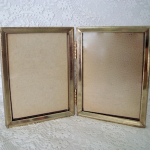 May include: A vintage, hinged double picture frame with a gold-colored metal frame. The frame is open, revealing two beige-colored backing boards. The frame is resting on a white lace surface.