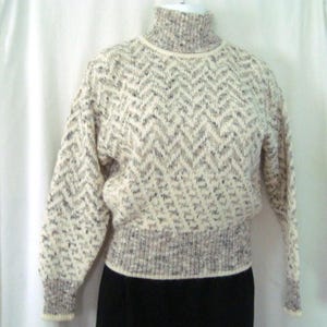 Könnte beinhalten: Ein langärmeliger Rollkragenpullover mit cremefarbener Basis und einem sich wiederholenden Zickzackmuster in Braun-, Grau- und Schwarztönen. Das Kleidungsstück verfügt über einen gerippten Kragen, Bündchen und einen breiten Saum in dunklerer Strickoptik.