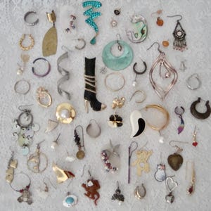 May include: A large collection of assorted jewelry pieces including various earrings, hoops, dangles, and pins. The items feature diverse shapes, colors, and materials such as metal, plastic, and beads, displayed in a scattered arrangement on a white lace surface.
