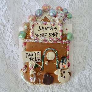 May include: A gingerbread house ornament featuring a sign that reads SANTA'S WORK SHOP and a smaller sign reading NORTH POLE. The structure is decorated with colourful glittery spheres, a snowman figure, and a gingerbread person figure.