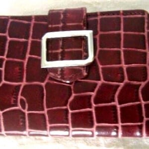 May include: A rectangular wallet in a deep burgundy color with a glossy finish and a textured crocodile skin pattern. The wallet features a central vertical strap secured by a polished silver-toned rectangular metal buckle centered on the front.