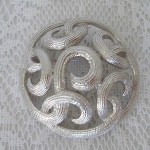 Puede incluir: Broche plateado con diseño circular. El broche presenta una serie de formas onduladas texturizadas dentro de un marco circular. La superficie texturizada captura la luz, creando reflejos y sombras. Un accesorio clásico.