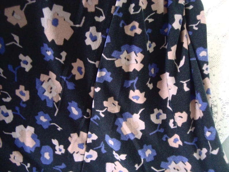 May include: Close-up of a dark fabric with a floral pattern. The design features small, stylized flowers in shades of light pink and blue against a dark background. The fabric appears to be soft and flowing, suitable for clothing.
