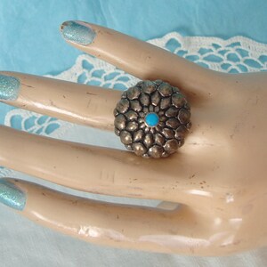 May include: A silver-toned ring featuring a circular, textured, petal-like design arranged in concentric layers. The center of the ring is accented with a small, round, bright turquoise stone. The metal surface has a darkened, oxidized finish.