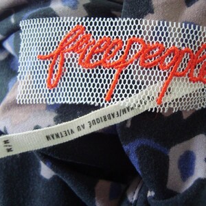 May include: A close-up shot of a clothing label featuring the brand name "free people" in orange embroidery on a white mesh. The label is sewn onto a fabric with a floral pattern in shades of blue, black, and tan. A smaller tag reads "M/M FABRIQUE AU VIETNAM".