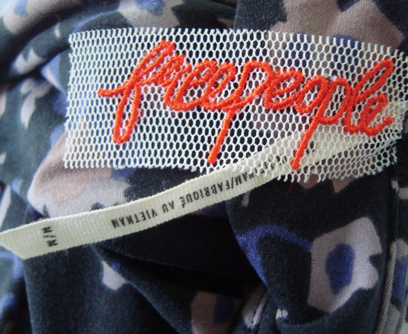 May include: A close-up shot of a clothing label featuring the brand name "free people" in orange embroidery on a white mesh. The label is sewn onto a fabric with a floral pattern in shades of blue, black, and tan. A smaller tag reads "M/M FABRIQUE AU VIETNAM".