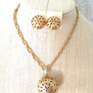 Puede incluir: Un conjunto de joyería en tono dorado que consta de un collar con un colgante redondo calado con un patrón radial y un par de pendientes redondos a juego. El collar presenta un diseño de cadena de eslabones texturizados en tono dorado.